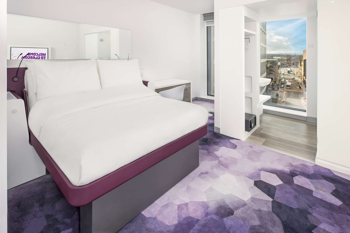 A modern bedroom with a large bed, a bedside table, a small desk, and a view of the city through a large window.