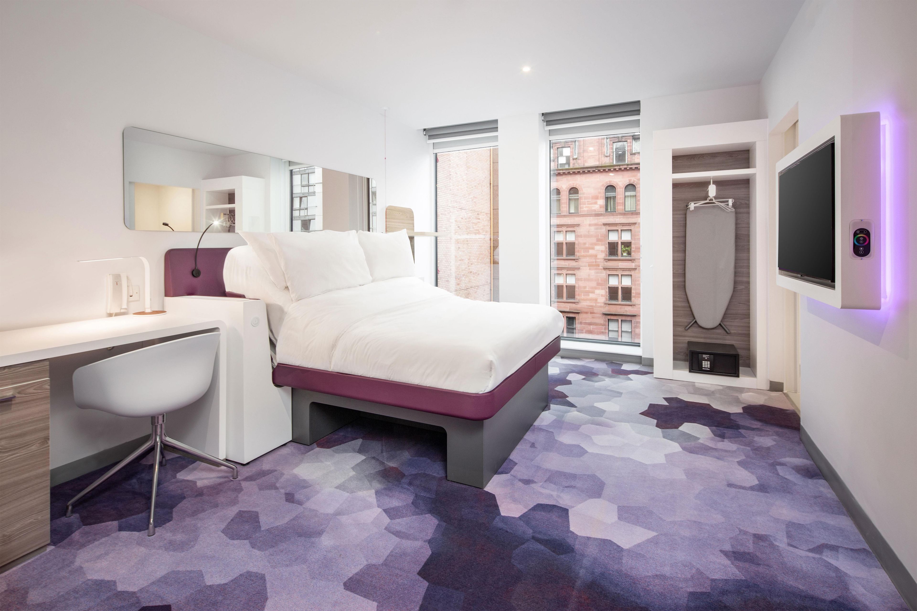 Una habitación de hotel moderna con cama, escritorio, silla y un gran ventanal con vistas a la ciudad.