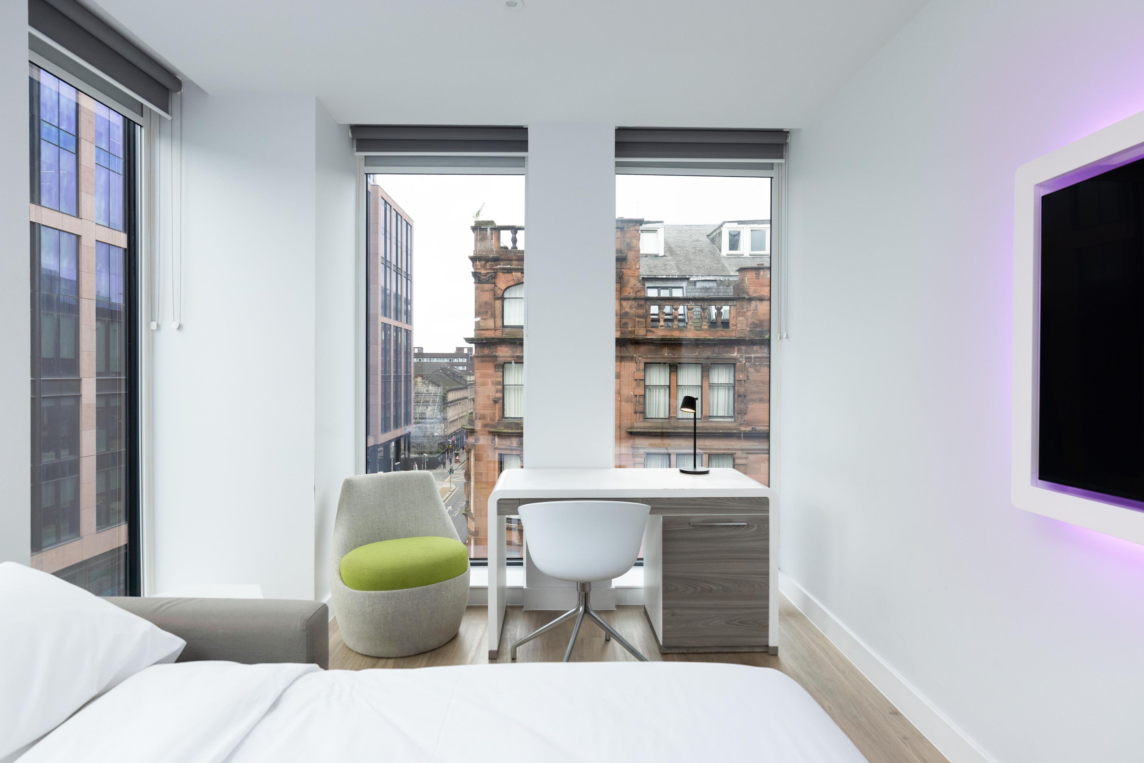Un dormitorio moderno con cama, escritorio y silla, con amplias ventanas y vistas a la ciudad.