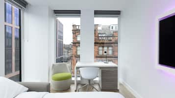 Un dormitorio moderno con cama, escritorio y silla, con amplias ventanas y vistas a la ciudad.