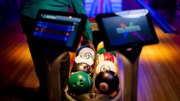 Una bolera con una persona con camisa verde a punto de lanzar, un rack de bolas y una pista de bolos con bolos.