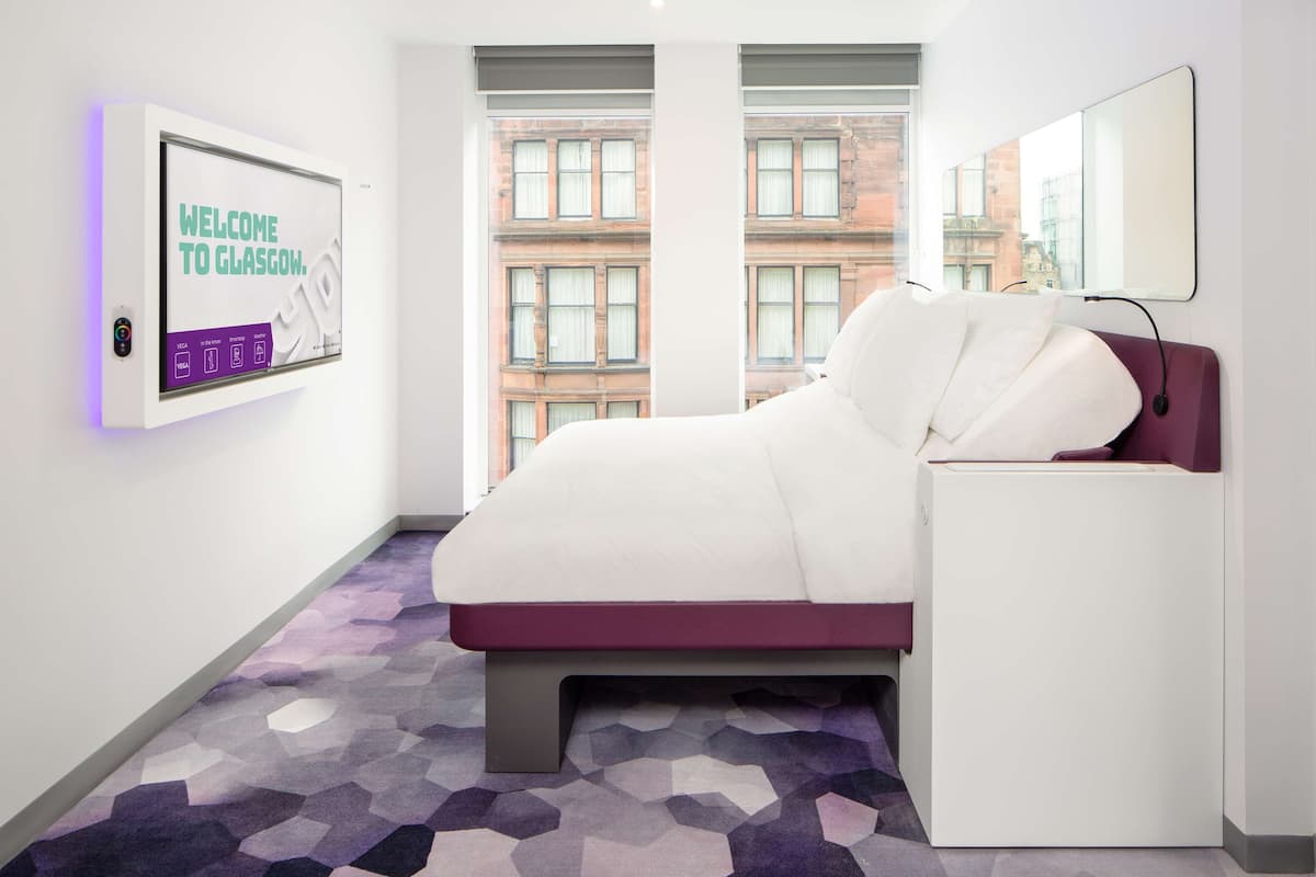 A modern hotel room with a large bed, a small bedside table, a wall-mounted TV, and a window with a city view.