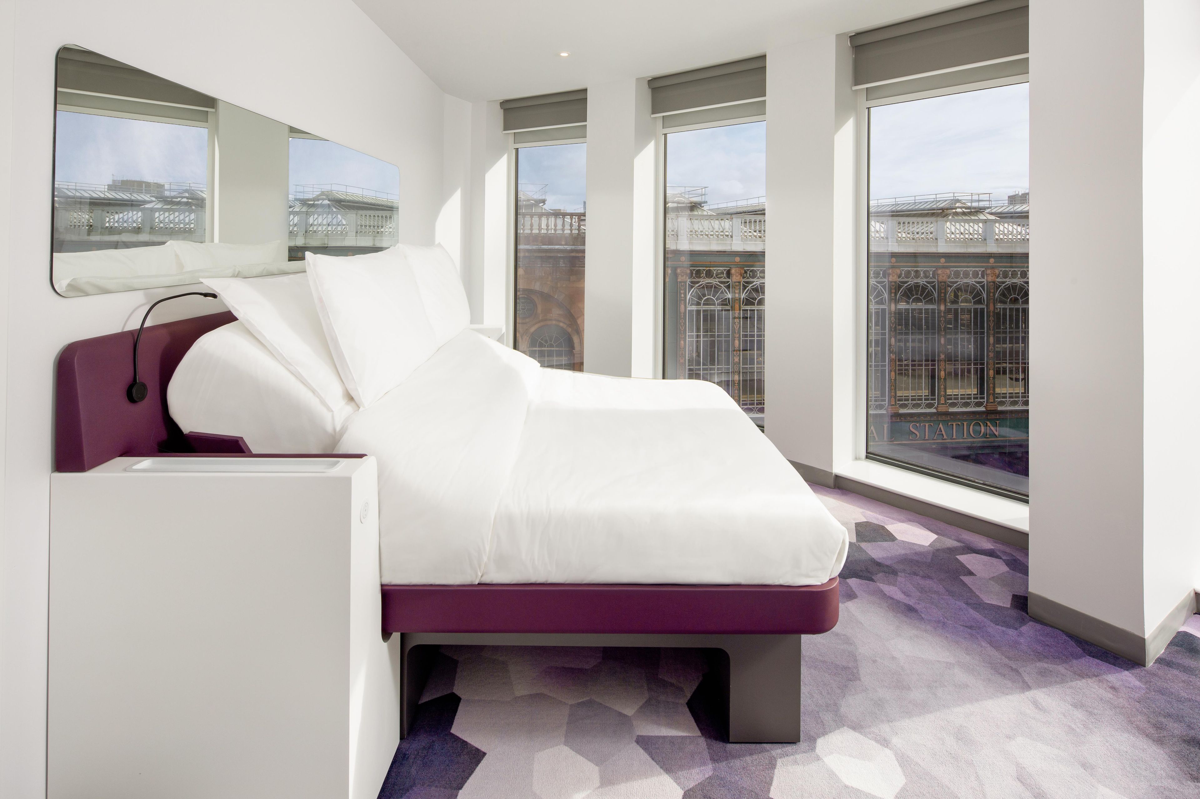 Habitación de hotel moderna con cama, escritorio y ventanas amplias que ofrecen vistas de la ciudad.