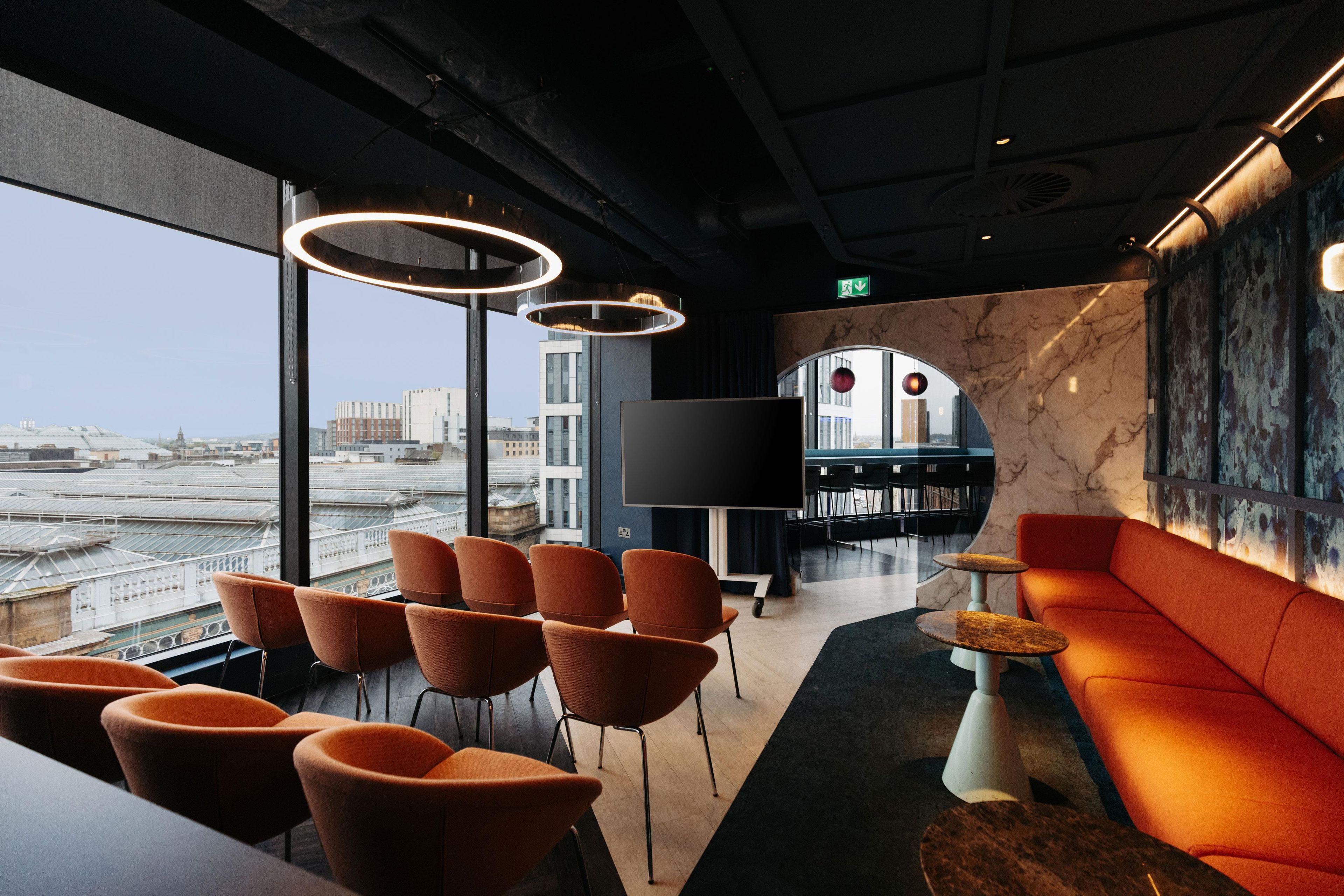 Een moderne lounge met uitzicht over de stad, met oranje zitplaatsen, een kleine tafel en een grote flatscreen-tv.