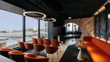 Een moderne lounge met uitzicht over de stad, met oranje zitplaatsen, een kleine tafel en een grote flatscreen-tv.