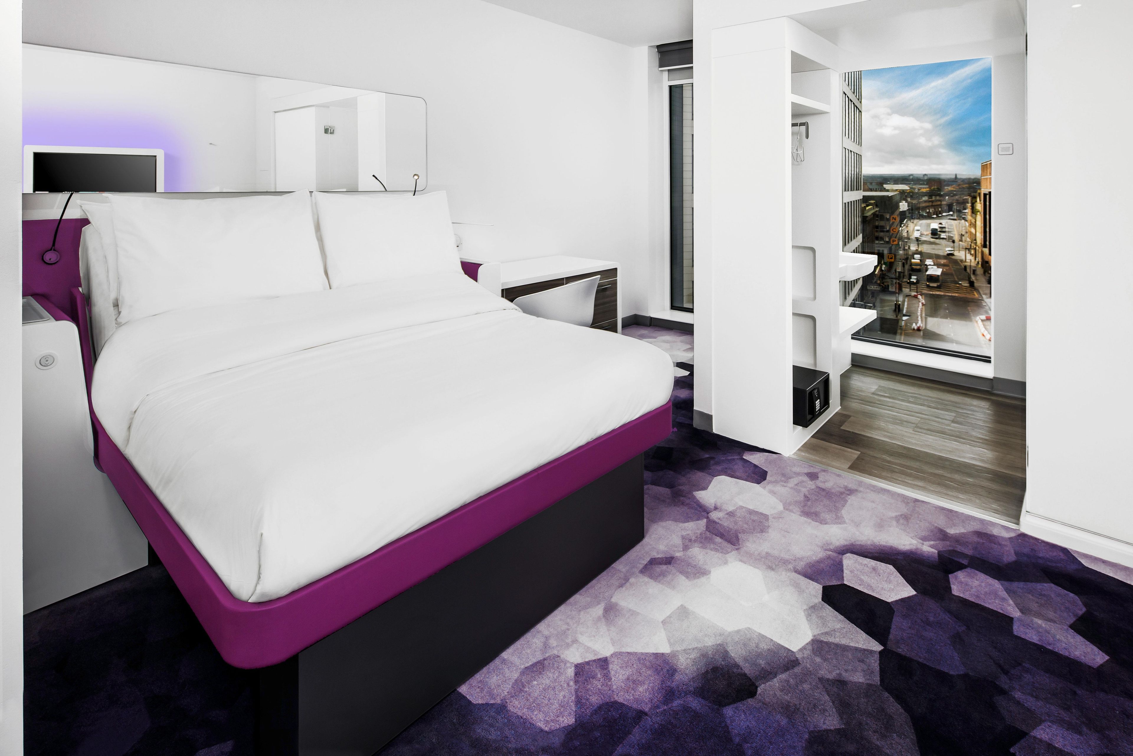 A modern hotel room with a large bed, bedside tables, a desk, and a city view through the window.
