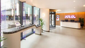 Zona de recepción de hotel con diseño moderno, un mostrador de recepción, un panel informativo digital y un amplio ventanal con vistas a la calle.