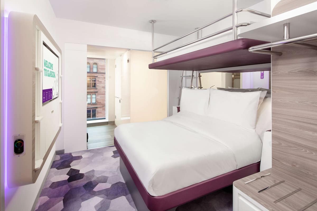 A modern hotel room with a bunk bed, a digital lock, a patterned carpet, and a view of the building's exterior.