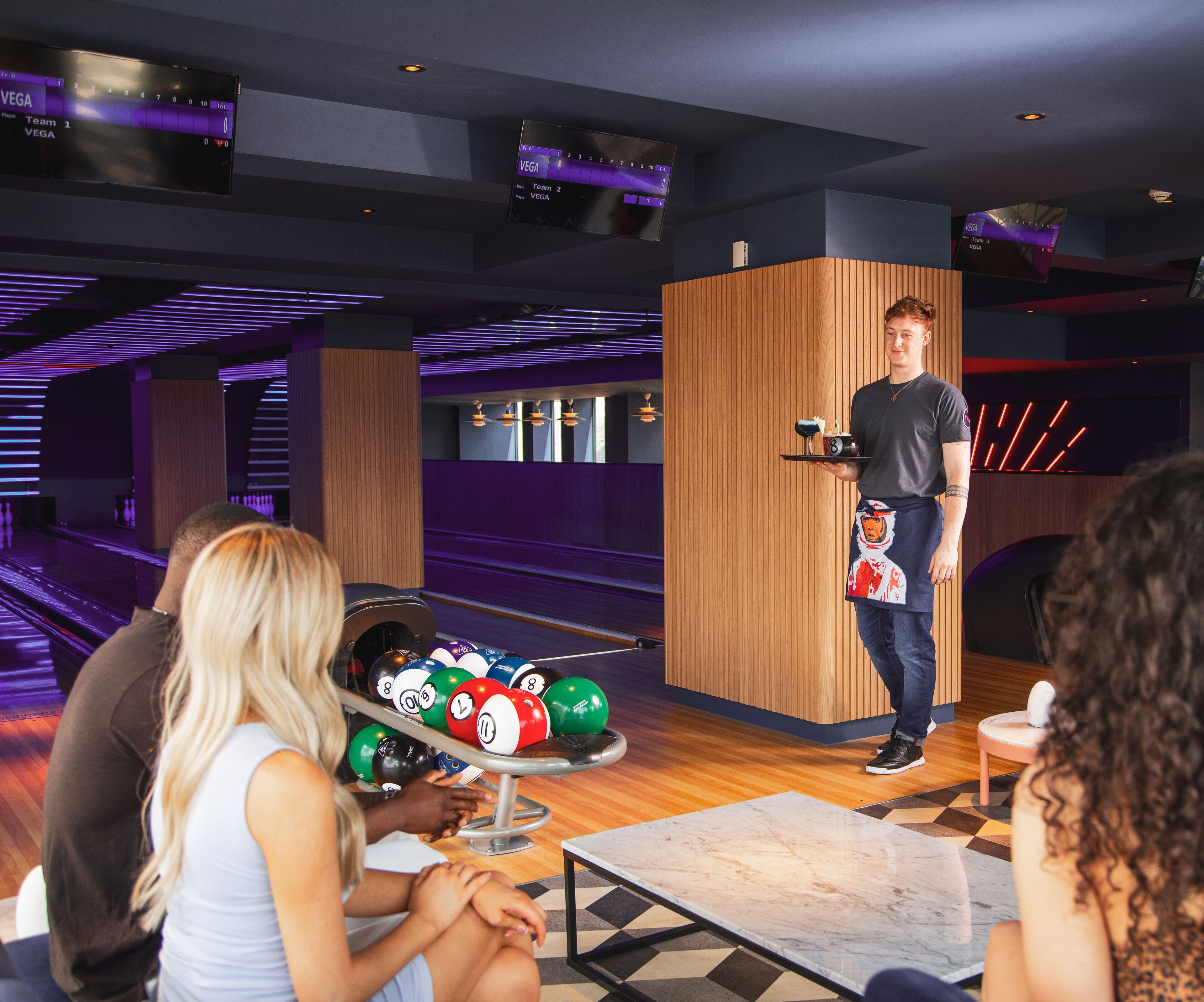 A bowling alley with a wooden counter, a scoreboard, and a table with bowling balls.
