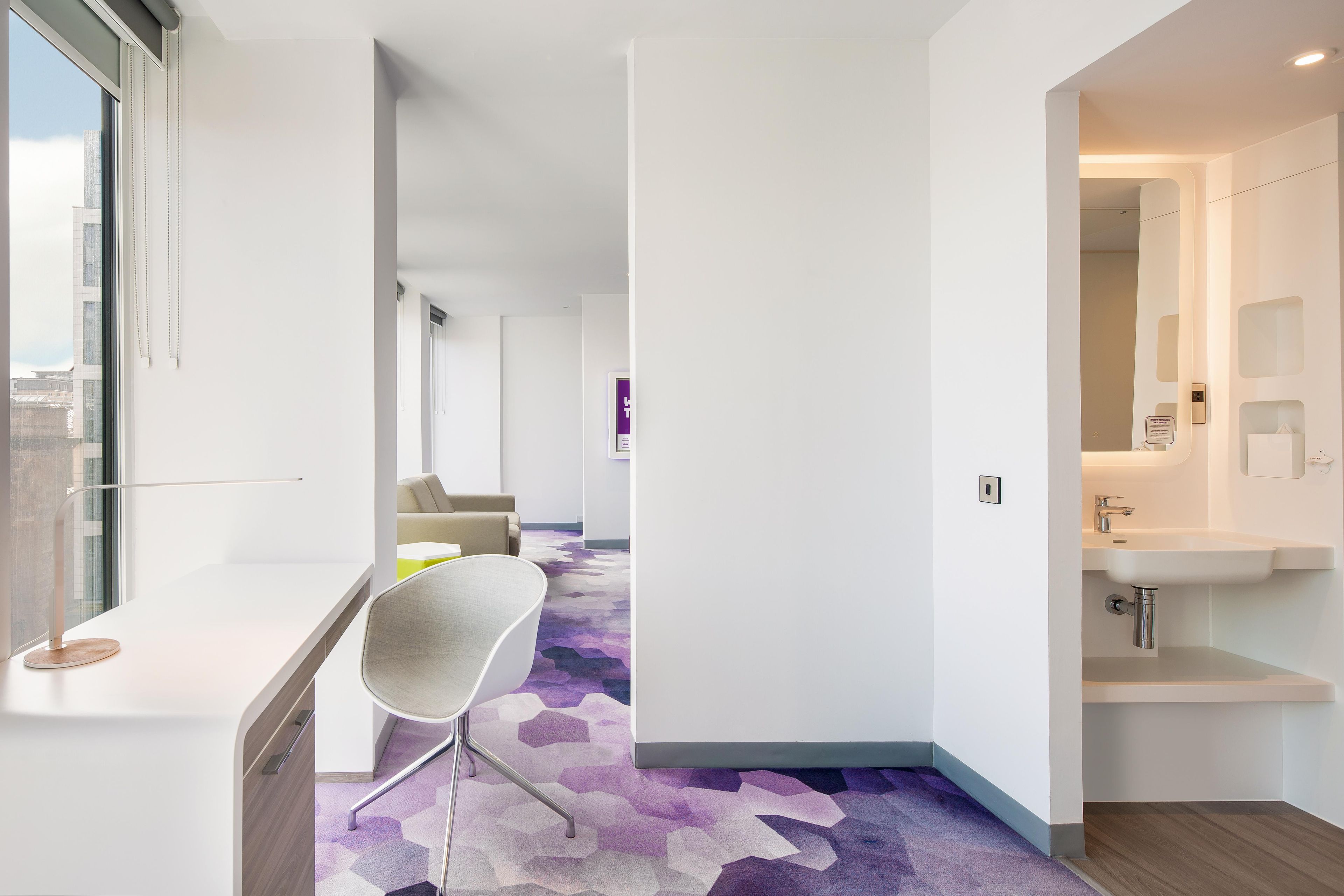 Una habitación de hotel moderna con un ventanal grande, un escritorio, una silla, un sofá y un baño con lavamanos y espejo.
