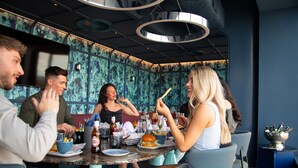 Un grupo de personas cenando en un restaurante con una decoración moderna, que incluye una mesa redonda con comida y bebidas, y una pared con un diseño original.