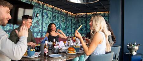 A group of people dining at a restaurant with a modern interior, including a round table with food and drinks, and a wall with a unique pattern.