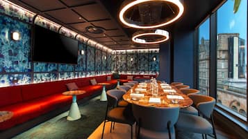 A modern lounge area with a long table set for dining, red seating, and a city view through large windows.
