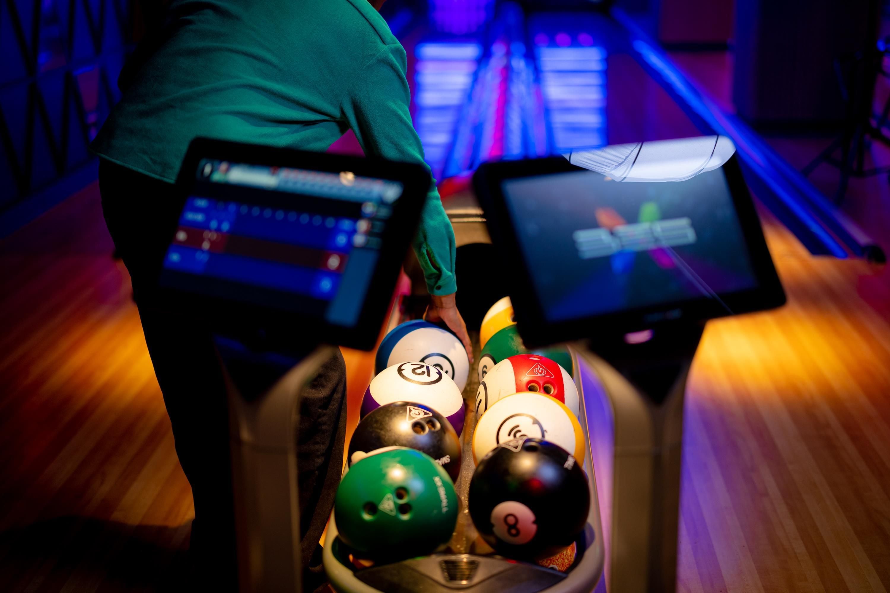 A bowling alley with a person in a green shirt preparing to bowl, a bowling ball rack, and a bowling lane with pins.