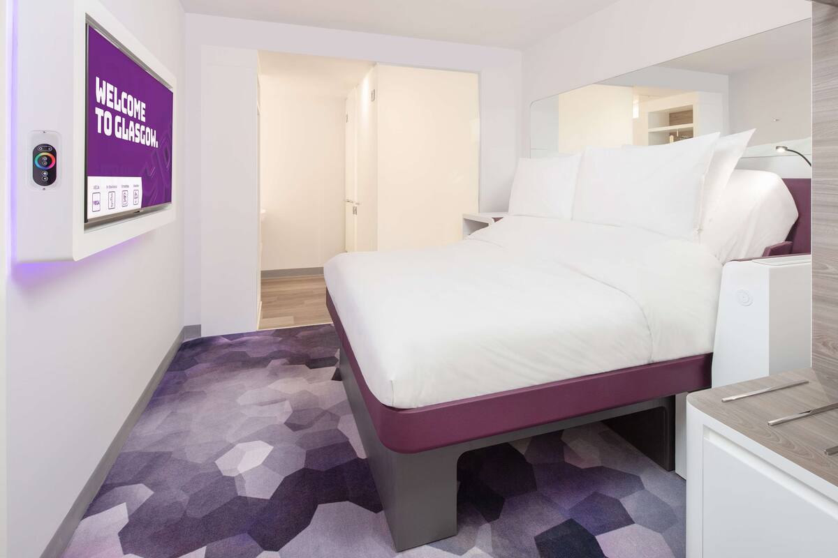 A modern hotel room with a bed, a bedside table, a mirror, and a wall-mounted digital clock.