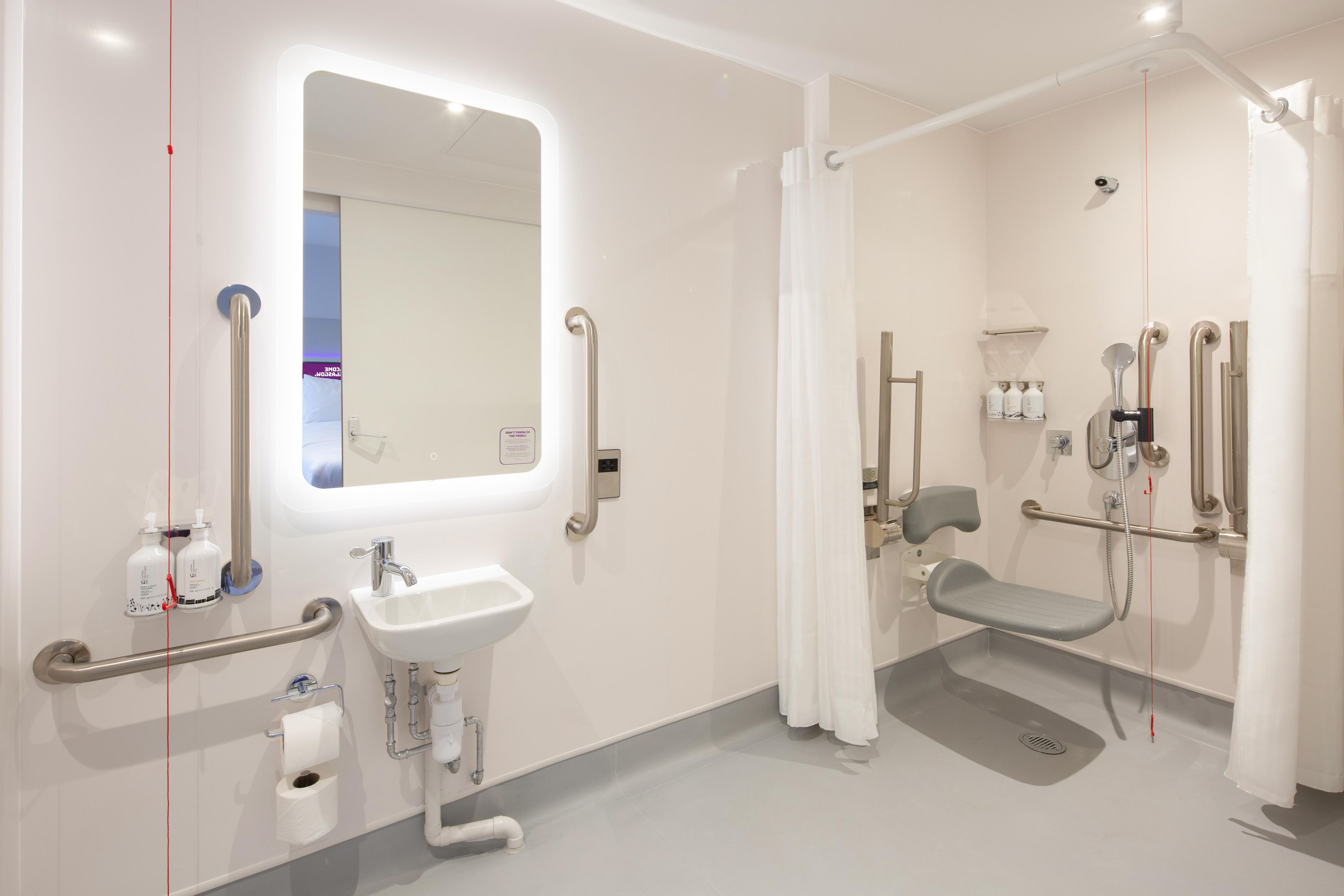 A modern bathroom with a sink, mirror, and shower area equipped with grab bars and a seat.