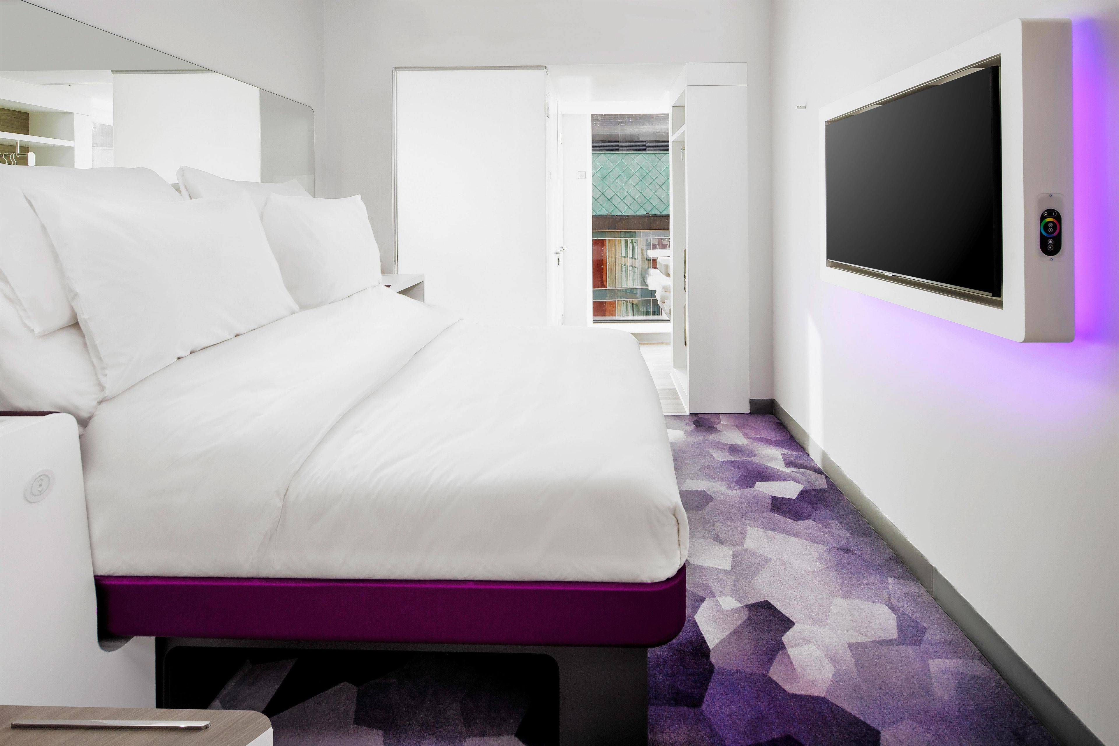 A modern hotel room with a bed, a TV mounted on the wall, and a purple carpet.