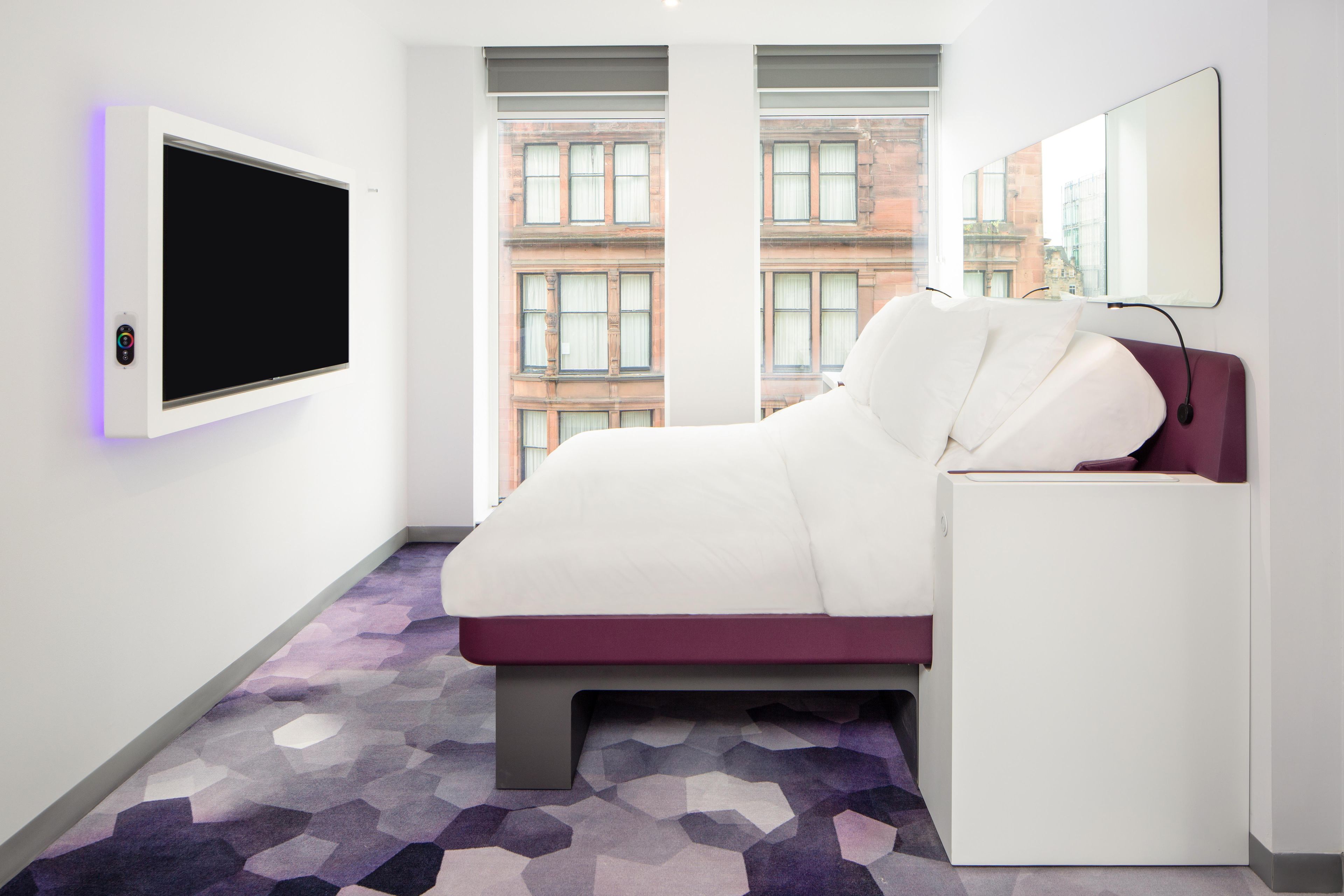 A modern hotel room with a large bed, a flat-screen TV mounted on the wall, and a window with a view of a building.