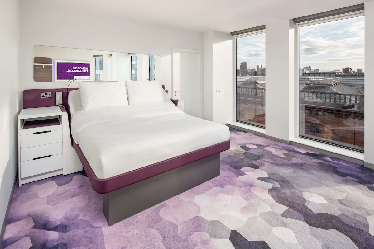 A modern hotel room with a large bed, a nightstand, a TV, and a city view through large windows.