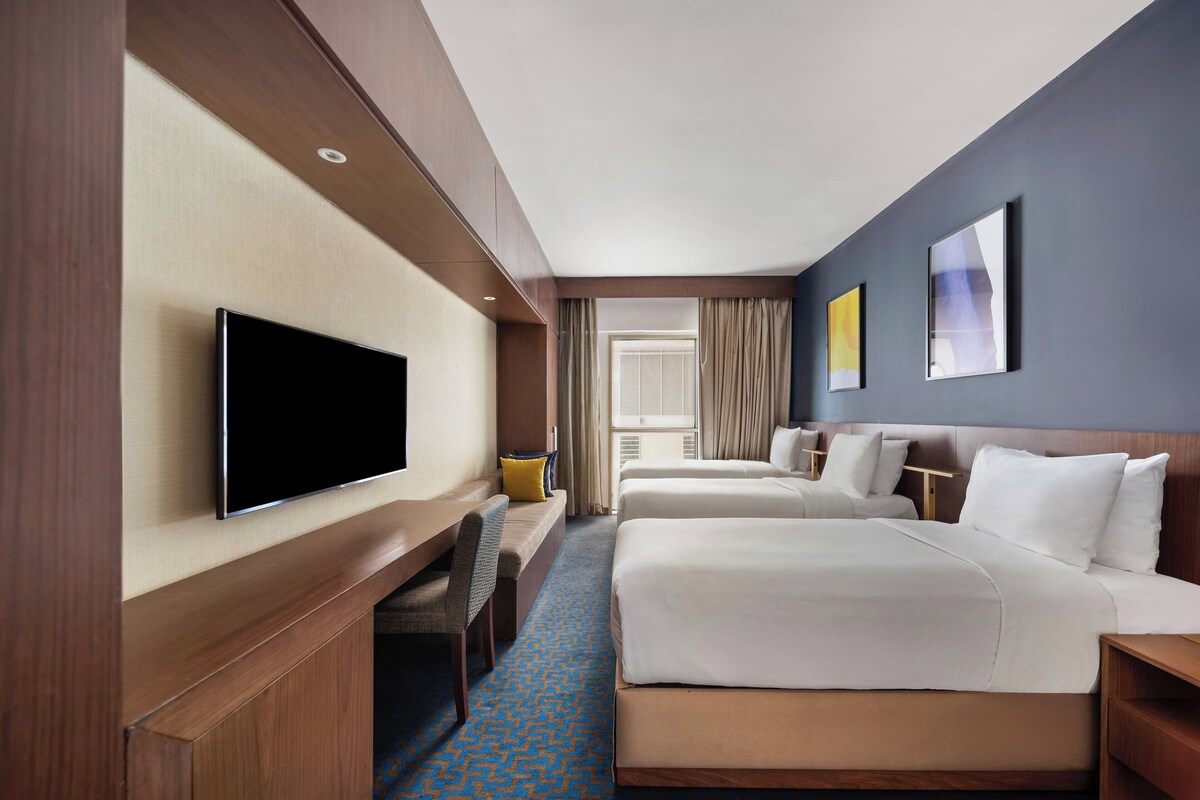 A hotel room with a flat-screen TV, two beds, a desk, and a window with curtains.