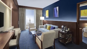 Habitación de hotel con sofá, sillones, una mesa de centro, una mesita de noche con lámpara y vistas a un paisaje urbano a través de amplios ventanales.