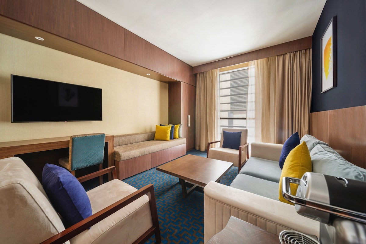 A hotel room with a flat-screen TV, a sofa, two armchairs, a coffee table, and a luggage cart.
