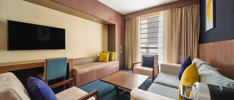 A hotel room with a flat-screen TV, a sofa, two armchairs, a coffee table, and a luggage cart.