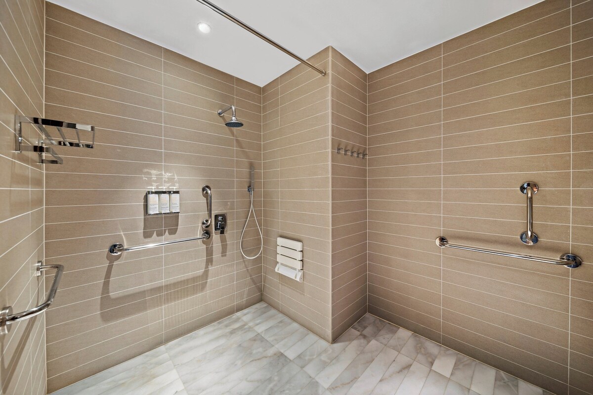 A modern bathroom with beige tiles, a shower with a handheld showerhead, a handheld showerhead, a towel rack, and a grab bar.