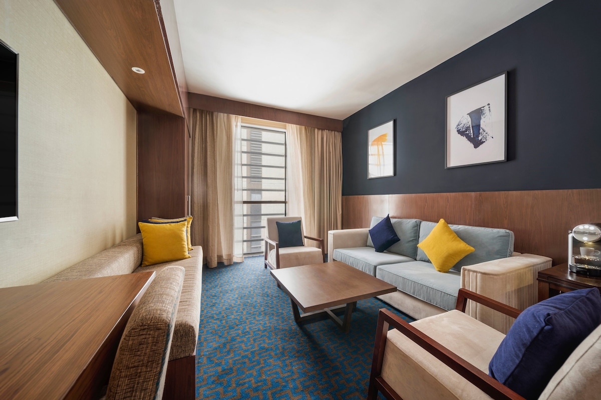 A hotel room with a sofa, armchair, and a small table. There is a window with curtains, a wall-mounted picture, and a carpeted floor.