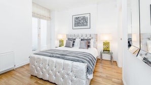 Une chambre à coucher avec un grand lit, deux tables de chevet, un lustre, un tableau encadré au mur et une fenêtre avec des stores.