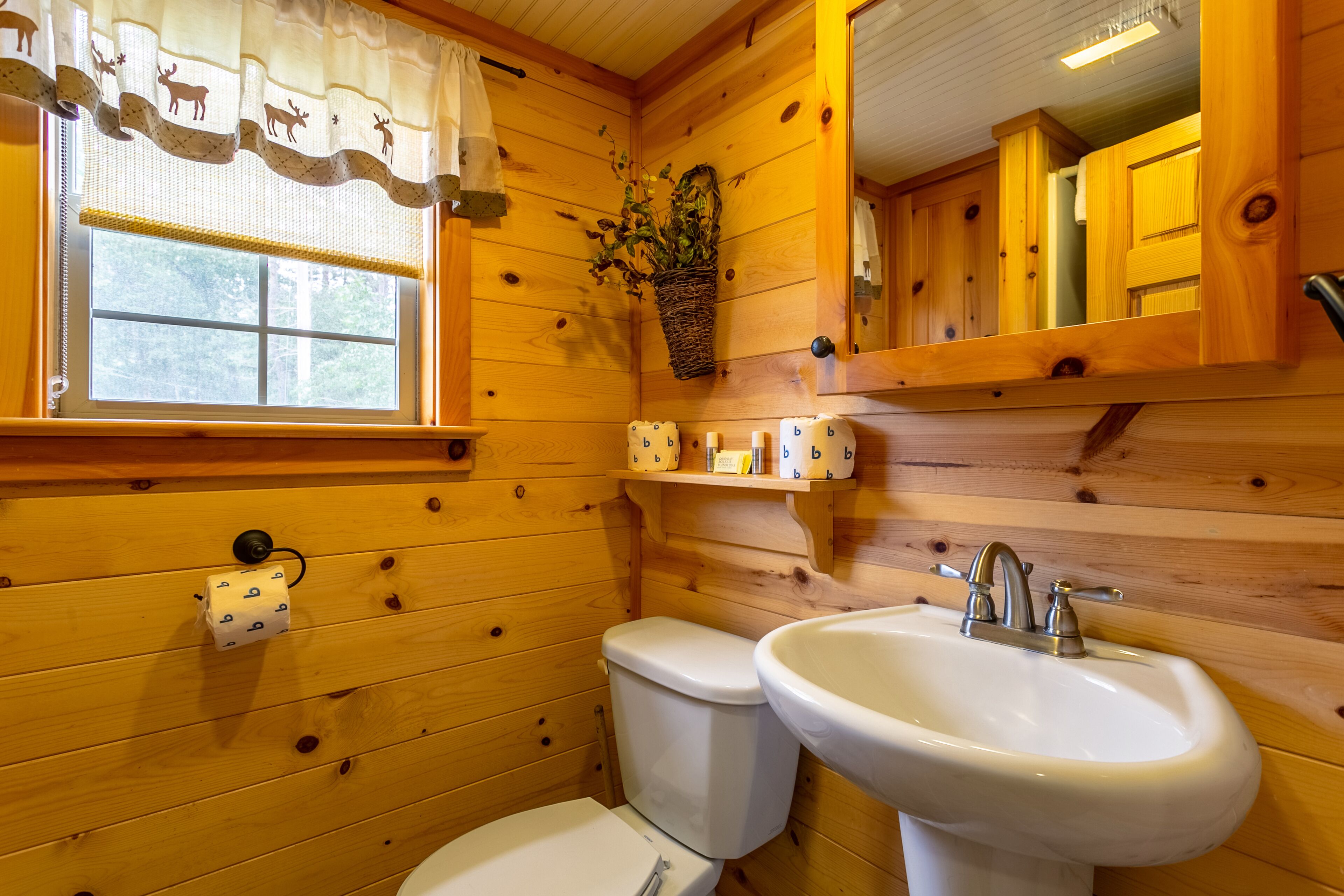 A wooden cabin bathroom with a toilet, sink, and window with a curtain.