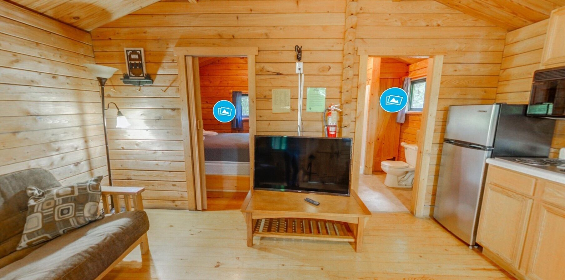 A wooden cabin interior with a sofa, a television, a kitchen area, and a bathroom visible through an open door.