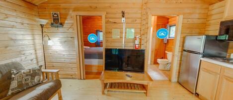 A wooden cabin interior with a sofa, a television, a kitchen area, and a bathroom visible through an open door.