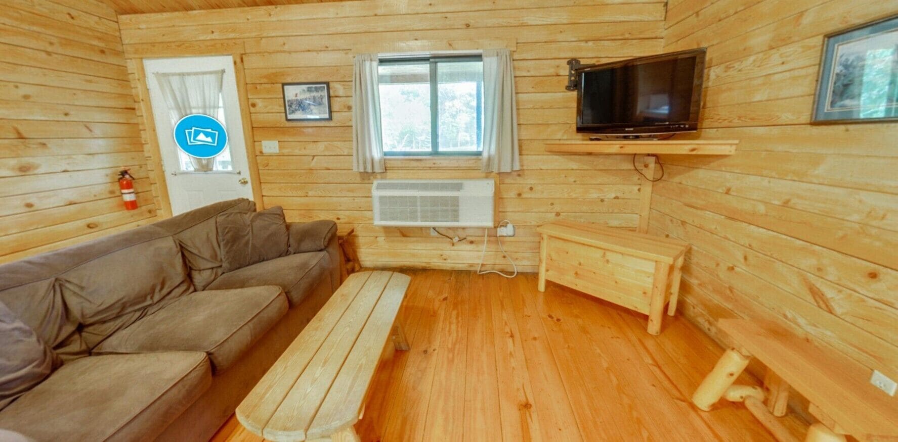 A log cabin interior with a sofa, a wooden bench, a TV mounted on a wall, and a window with curtains.