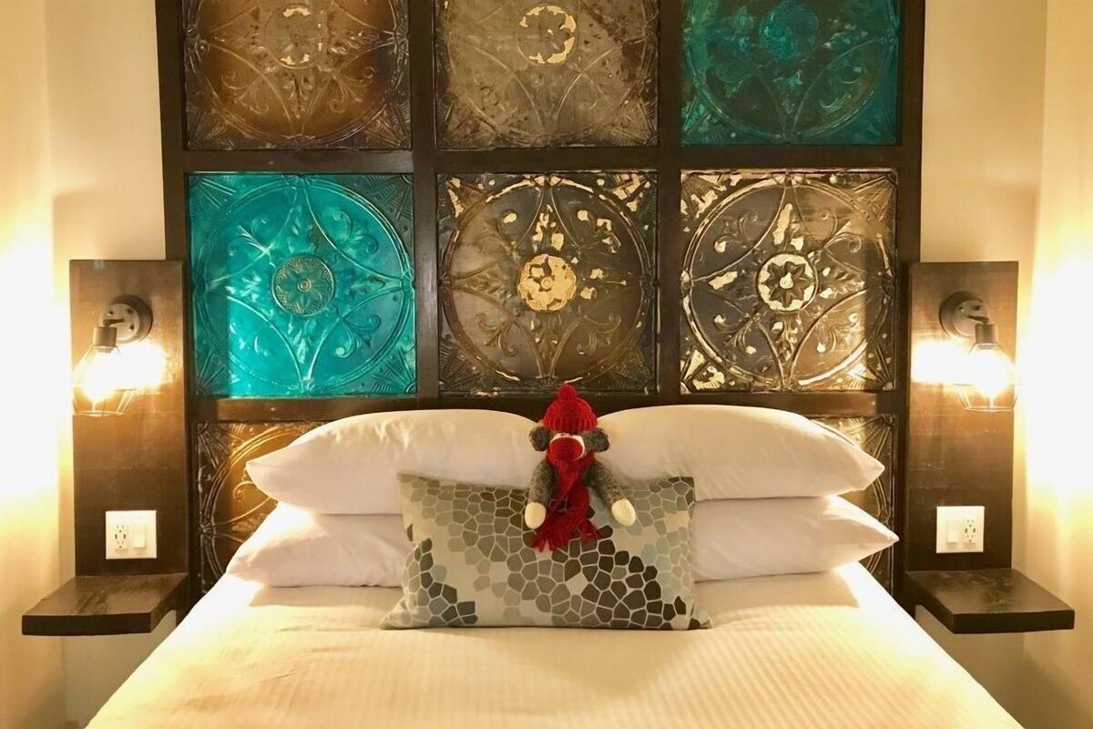A neatly made bed with a decorative pillow and a plush toy, flanked by two bedside tables with lamps, against a wall with a mosaic-style headboard.