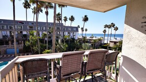 A balcony with wicker chairs and a pool view, overlooking a beach and palm trees.
