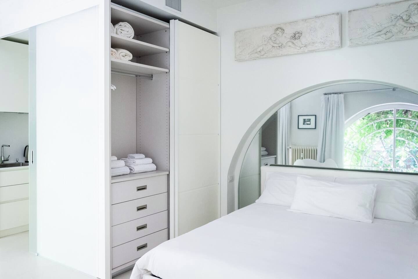 A modern bedroom with a large bed, a walk-in closet, and a view of greenery through a window.