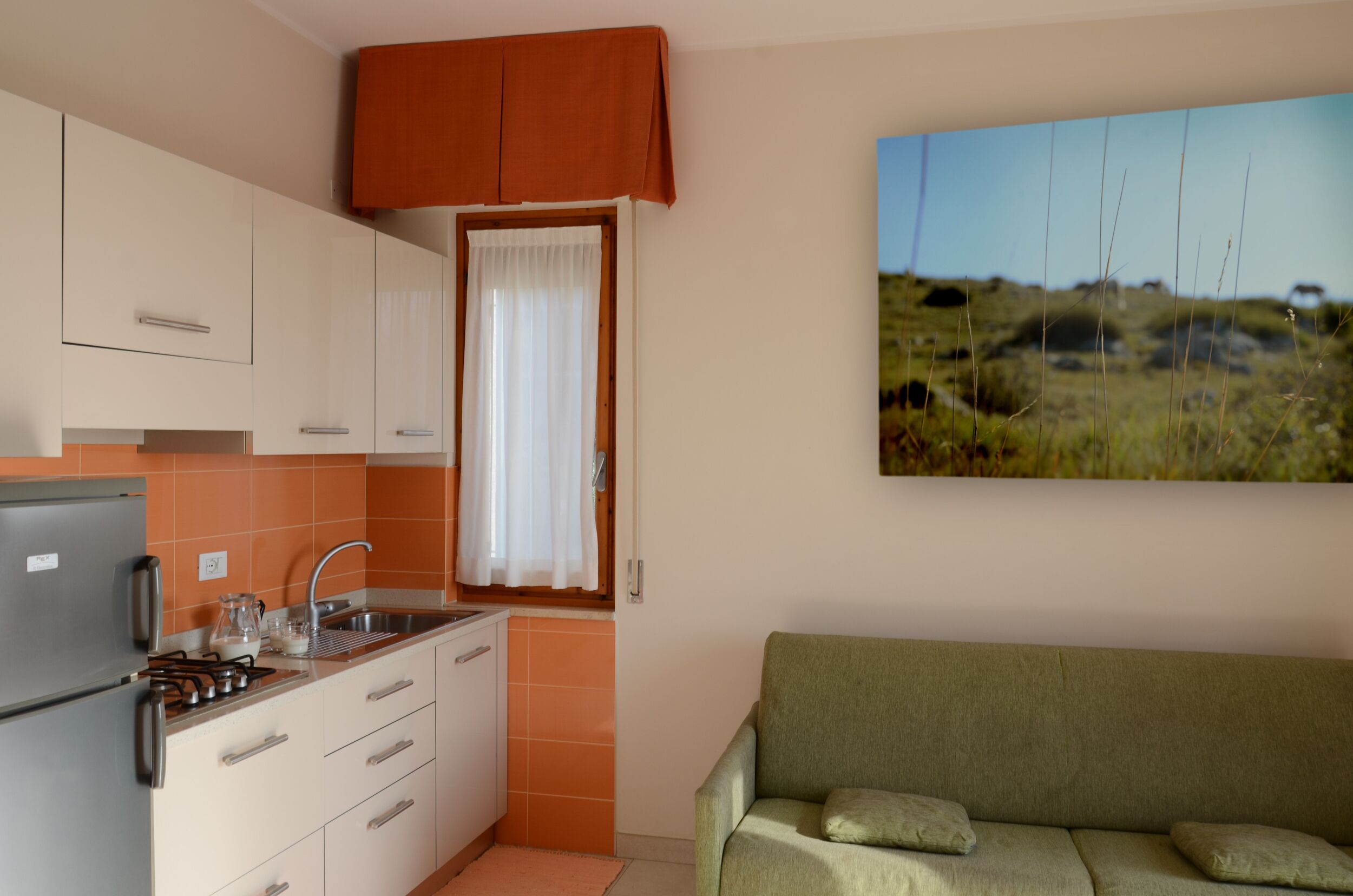 Une cuisine compacte avec des meubles blancs, un four encastré et un évier. On y trouve un canapé vert avec des coussins et un tableau représentant un paysage accroché au mur.