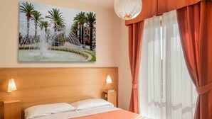 Ein Hotelzimmer mit einem Bett, einem Nachttisch, einer Wandleuchte, einem Fenster mit Vorhängen und einem Bild einer Fontäne.