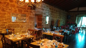 A rustic dining area with stone walls, wooden tables, and chairs. There are multiple dining tables set with plates, cups, and cutlery. A chandelier hangs from the ceiling, and a framed picture is visible on the wall. - Casa Rural Osante (Labastida)