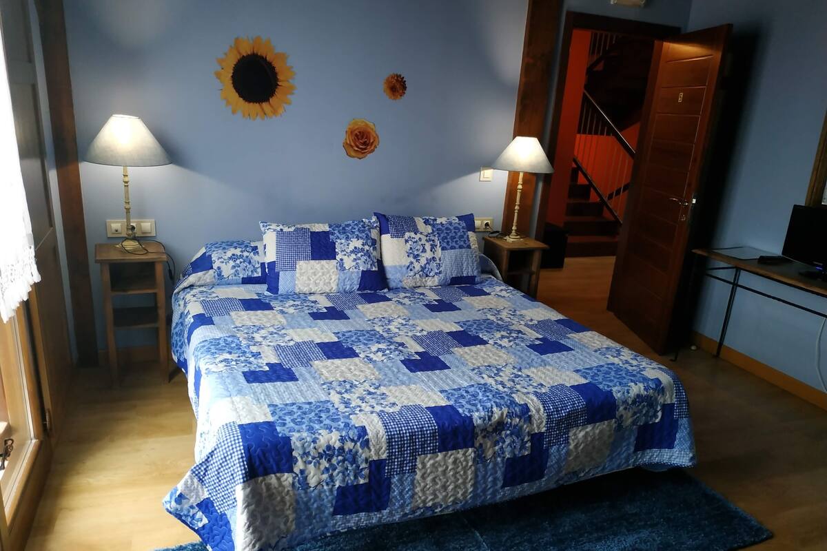 Una camera da letto con una coperta trapuntata blu e bianca, due abat-jour, un comodino in legno, un tavolino, un tappeto blu e un muro decorato con un girasole.