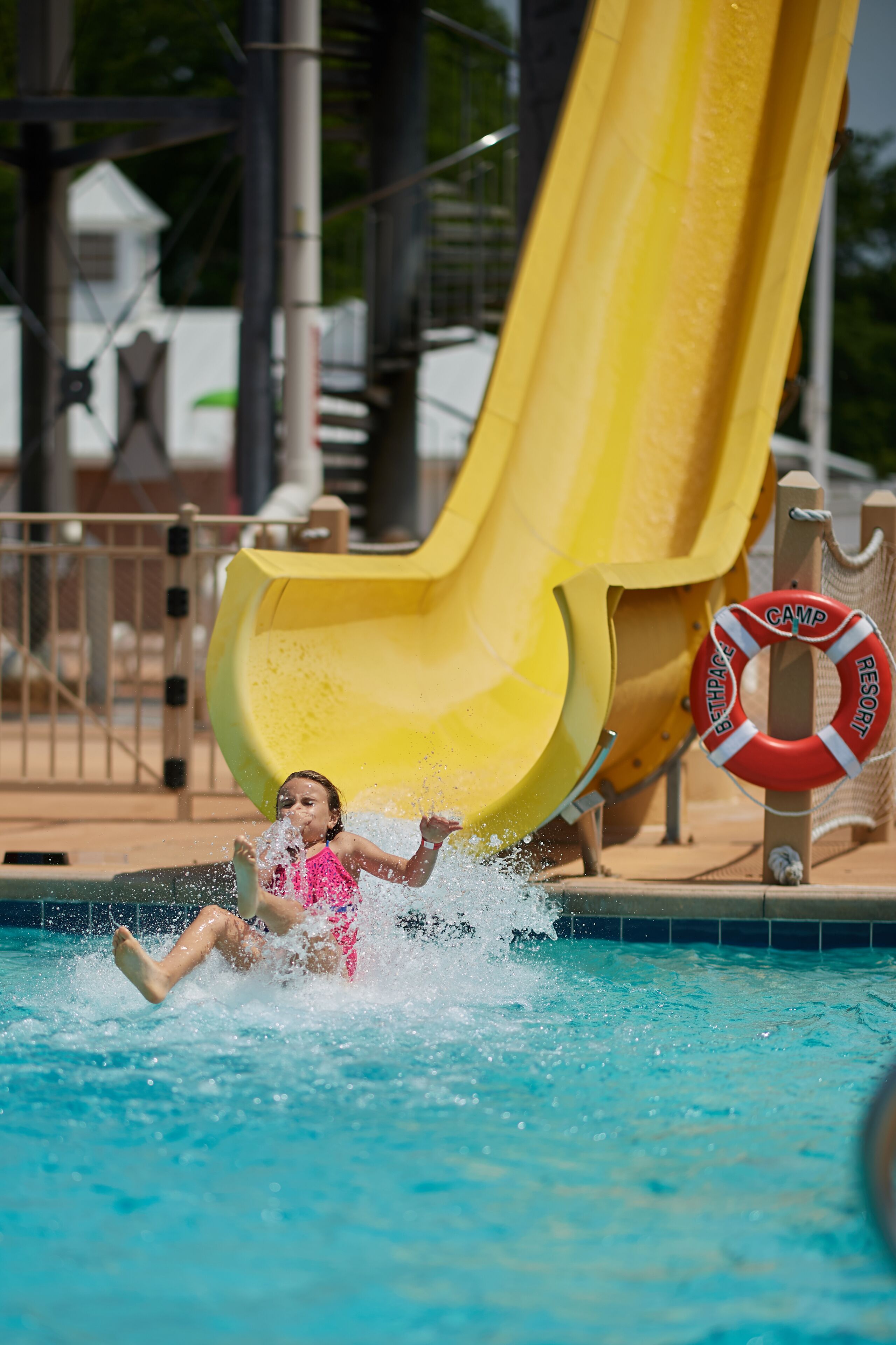 A child sliding into a pool with a yellow slide and a lifebuoy nearby.