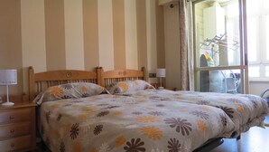 A double bed with floral bedding, wooden headboard, bedside tables with lamps, and a window with a view of outdoor equipment. - Front line with sea views garage + free wifi (Zarauz)