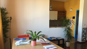 A room with a wooden table, books, a potted plant, and a kitchen in the background. - Casa Arcobaleno, an experience to try! (Firenze)