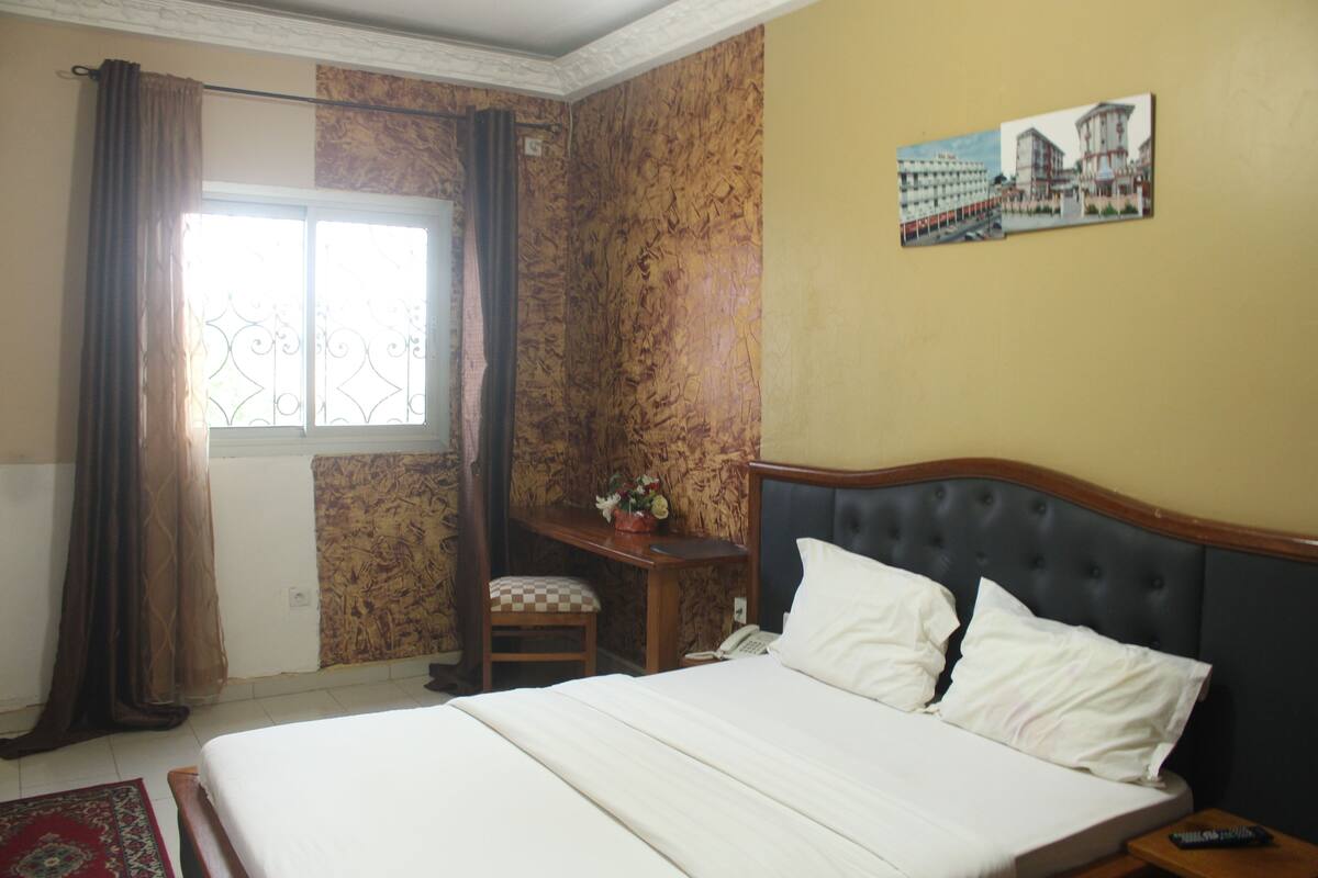 A hotel room with a large bed, a desk with a telephone, a chair, and a window with curtains.