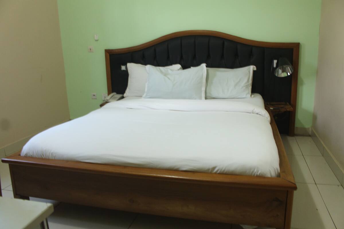 A standard hotel room with a wooden bed, white bedding, a nightstand with a lamp, and a plain wall.