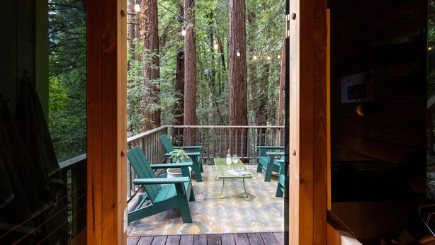 Una terraza de madera con sillas verdes y una mesa, con vista a un área boscosa.