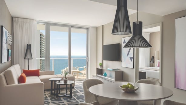 A modern living room with a sofa, dining table, and chairs, a large window with a view of the sea, and a balcony with outdoor furniture. - Hyde Beach House (Hollywood)