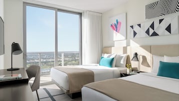 A hotel room with two beds, a desk, and a chair. There is a large window with a view of the city.