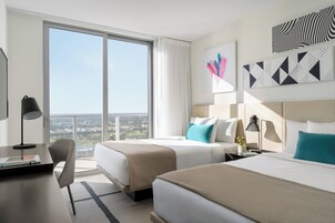 A hotel room with two beds, a desk, and a chair. There is a large window with a view of the city.